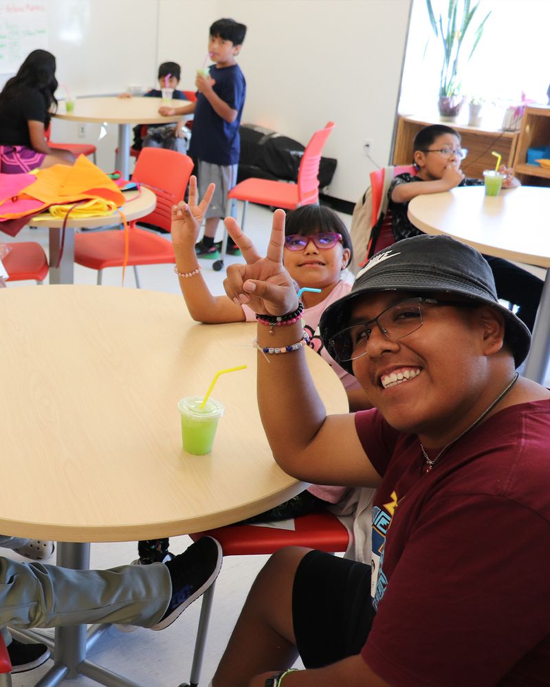 Picture of a young man in a classroom sitting at a table with a girl. Both the young man and the girl are giving a peace sign to the camera.