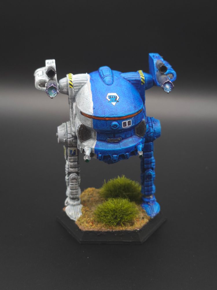 JagerMech III miniature painted blue on the left 2/3 and white on the right 1/3. For the 10th Lyran Guard. The silver piping detail divides the two colors vertically. It is displayed on a finished base with bushes.