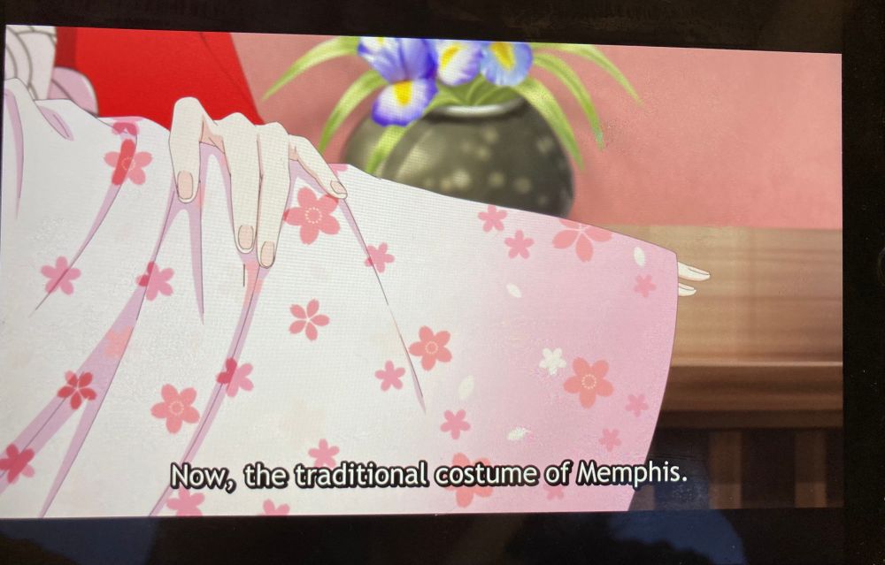 Phone “screenshot” from “May I Ask For One More Thing?” where a character is putting on a yukata with the subtitle saying “Now, the traditional costume of Memphis.”