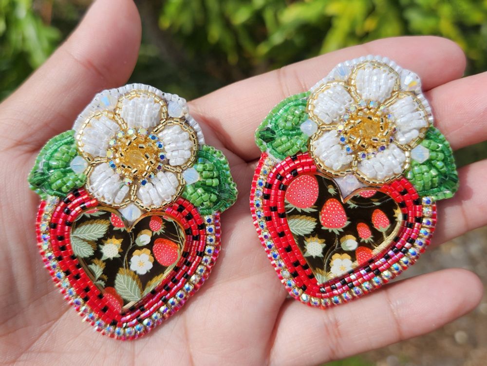 A pair of earrings. White raised beadwork flowers sit ontop of a golden mirror cab. The beads surrounding the can mimic the look of a strawberry. Made with 11/0 delica beads