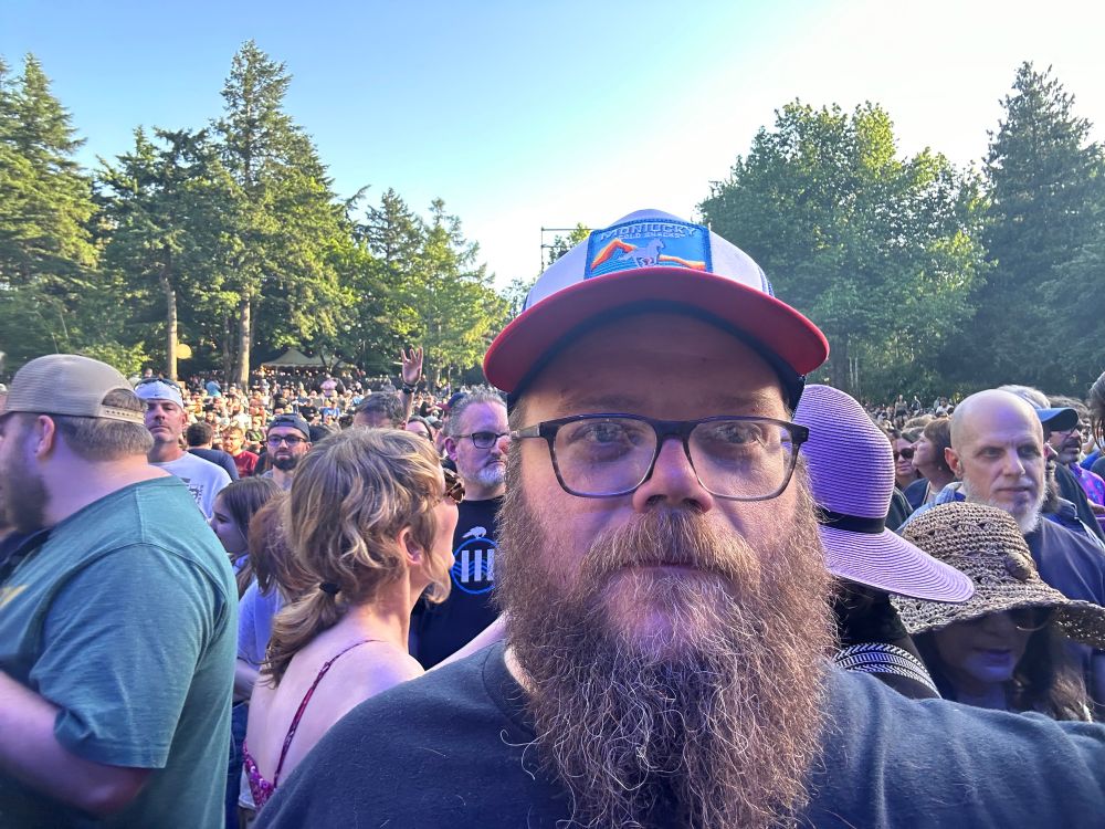 Selfie of a bearded white make in his early 40s. He is wearing black rimmed glasses and a Montaucky beer hat. Thousands of concert-goers spread out behind him. 
