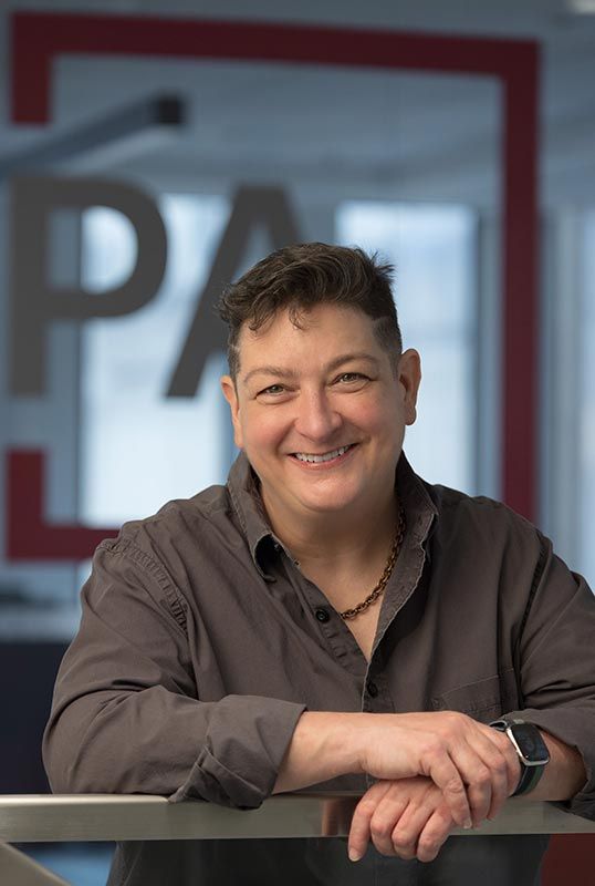 Photo of a non-binary person, smiling in a grey button-down shirt, holding their hands crossed and relaxed in front of them, resting on a metal railing. Their hair is cut in a fade w short sides and a line shaved on each side.