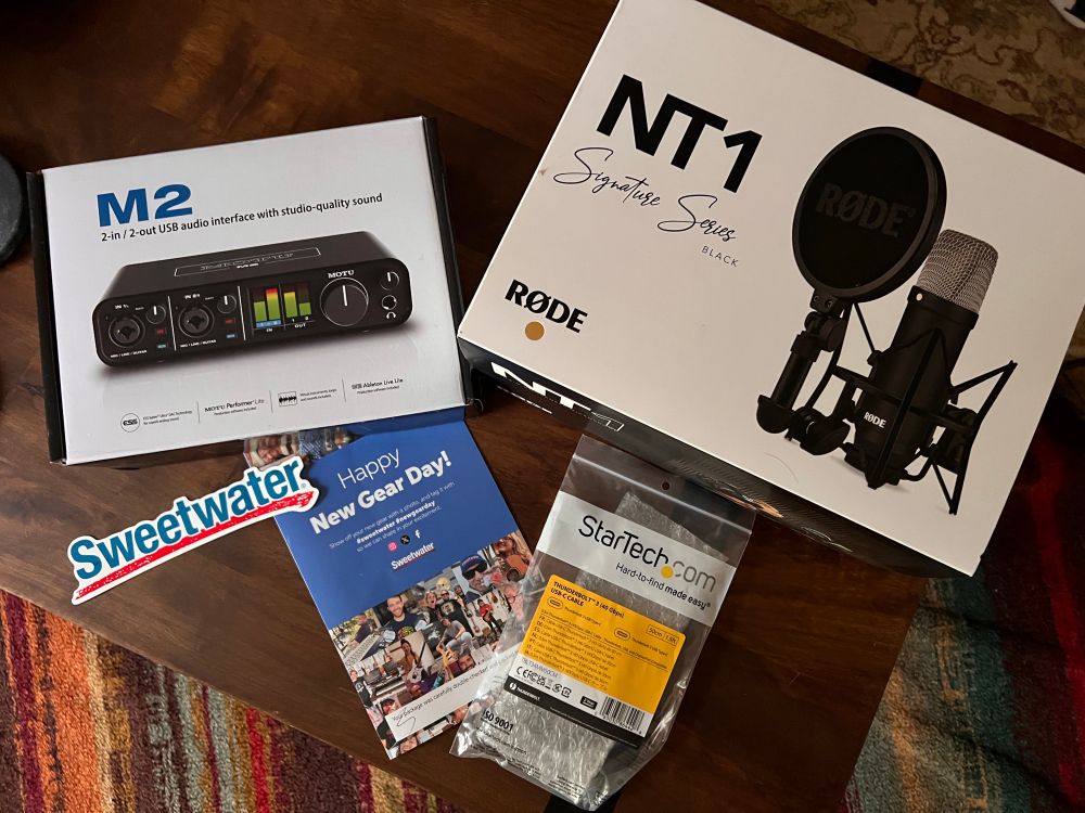 Overview pic of sound equipment—a Motu M2 audio interface box and a Rode NT1 microphone box—plus a Happy New Gear Day card, a Sweetwater sticker, and a cable in a bag. Not shown: the bag of candy they also sent me. 
