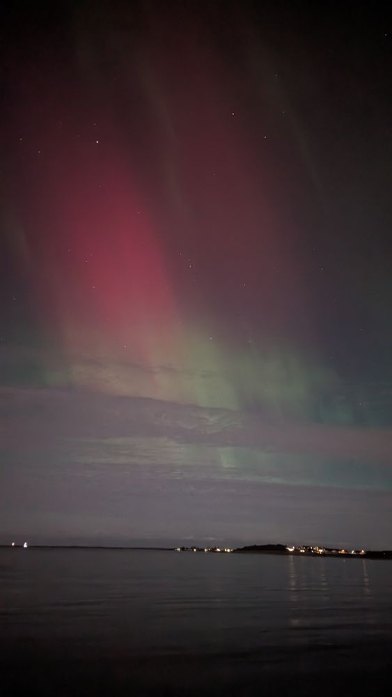 Red and green northern lights above the sea with some lights of houses at the horizon.