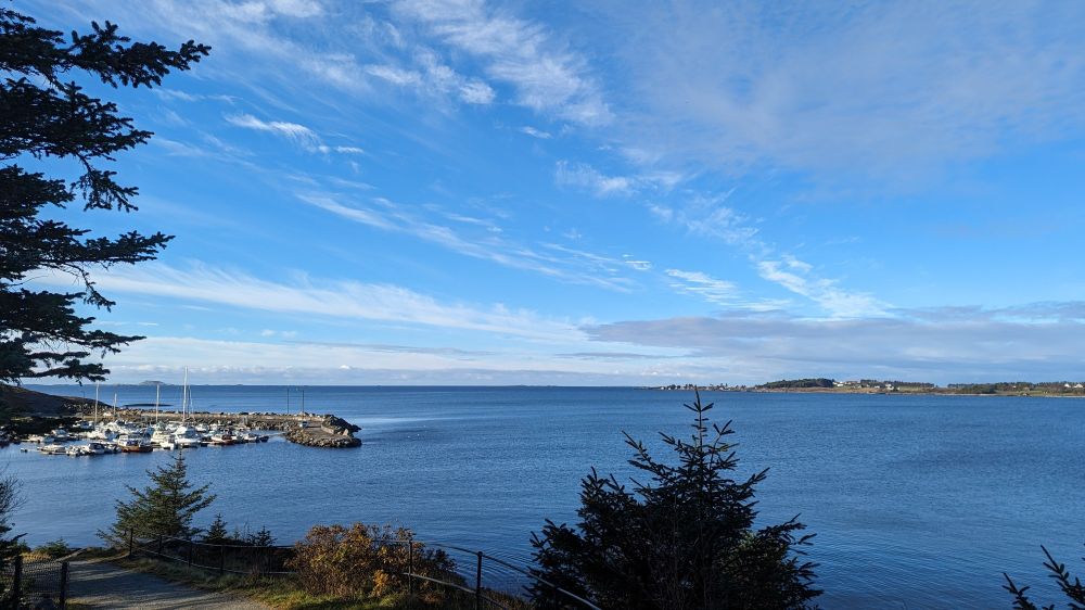 View over a calm sea from a slightly high position with a small harbour on the left and nearly clear blue sky above.
