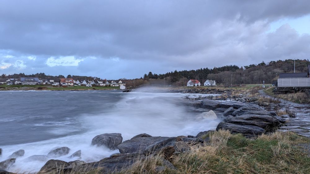 Stormy sea water spray over a breaker at the coast in Norway. Houses and trees at the distance.