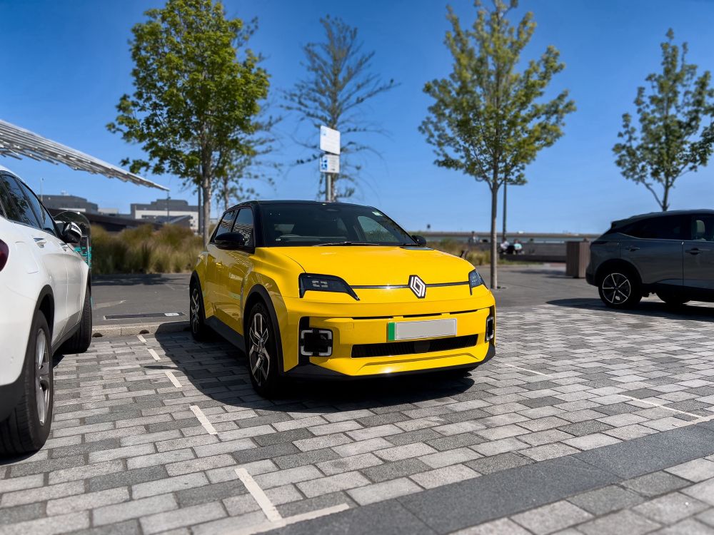 A front 3/4 photograph of a small compact car parked in a cobble stoned parking lot. The car has warm yellow paint with a black roof. 