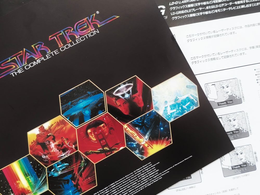 View from above of the included booklet, titled 'Star Trek - The Complete Collection', in its vintage and colorful font. A few hexagons can be seen underneath, each part of the poster for each movie, as well as a nice shot of the Enterprise from the rear.