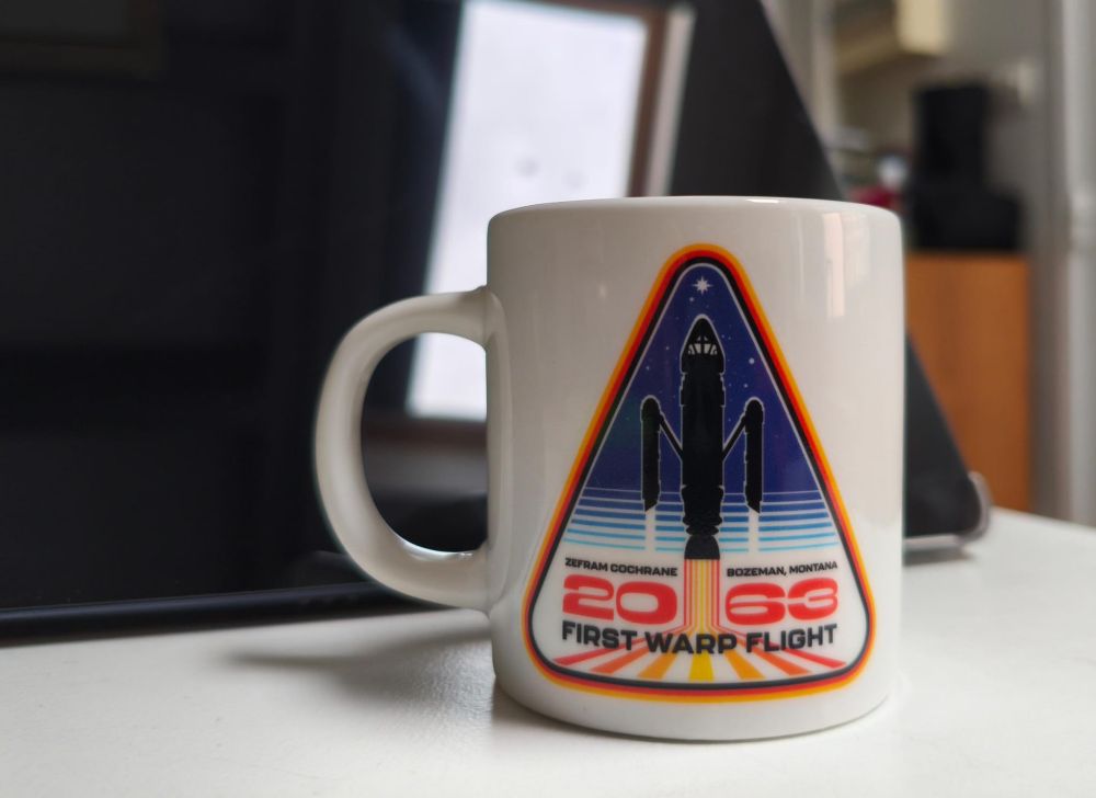Color picture featuring a white espresso cup with a triangular design, which encompasses Zefram Cochrane's vessel as a silhouette flying up under a vintage graded navy blue background with a few stars; a "2063" in red can be seen lower down the triangle, with the mention ''First Warp Flight'' right below in a black font. Such a possession might seem confusing for a tea drinker, but it is a piece of history this one is honored to have.