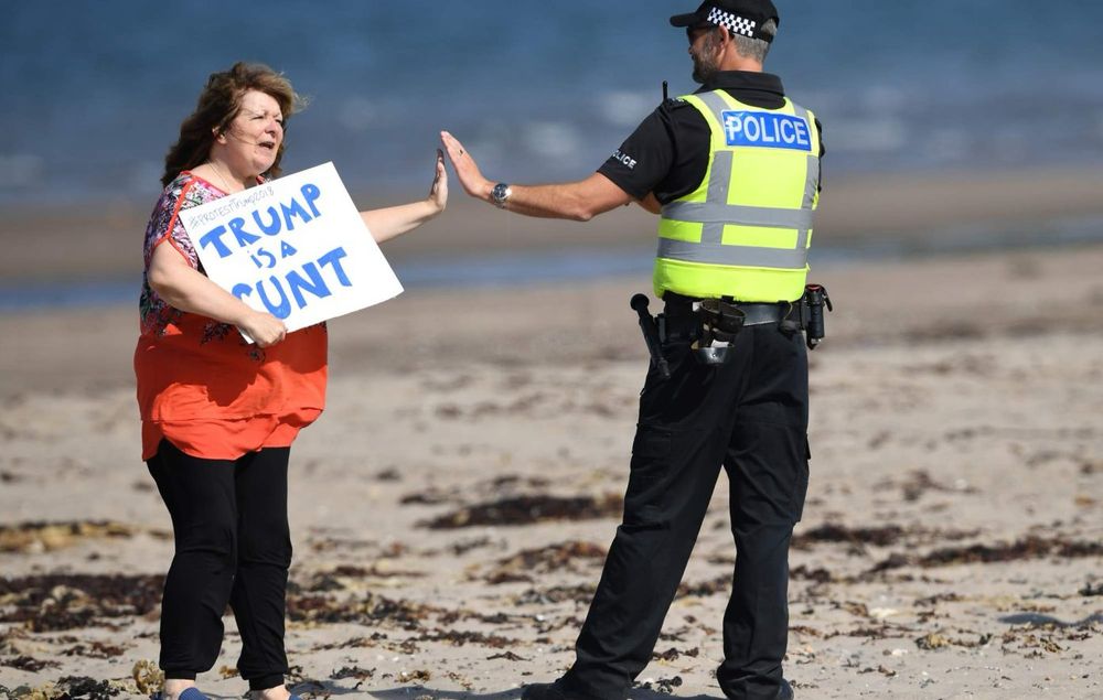 Janey Godley high fiving a cop and holding a sign that says tr@mp is a c@nt