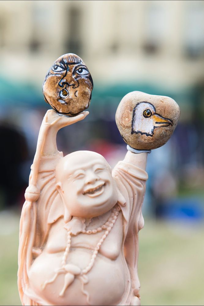 An image which shows a buddha statue holding up a stone in both hands - one with a human face and the other is the head of a seagull. 