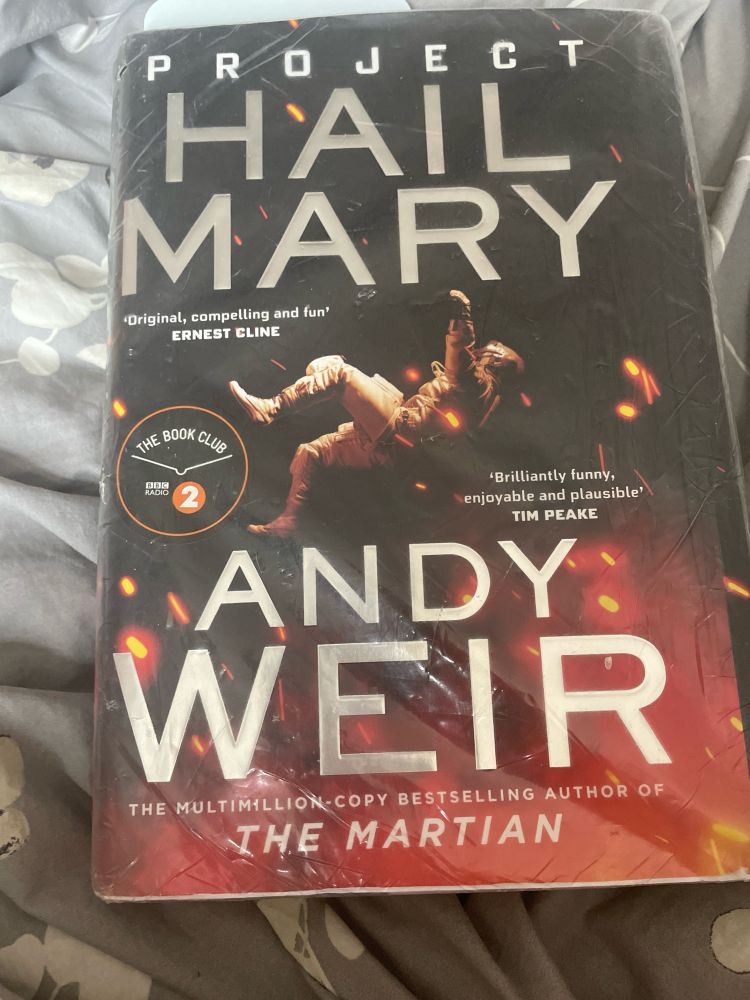 A hardback copy of Project Hail Mary by Andy Weir, which has an astronaut floating (flailing?) across a dark sky with red and yellow flashes of light.