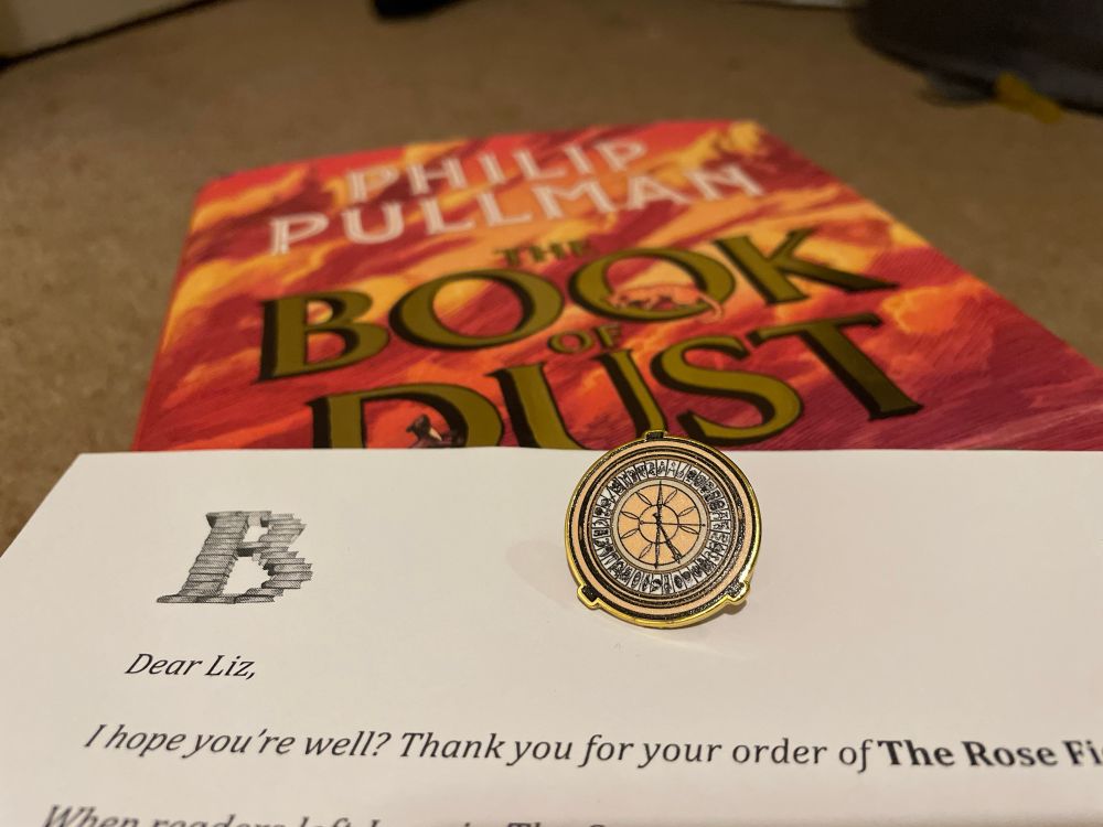 A shiny gold alethiometer pin badge on top of a hardback copy of The Rose Field by Philip Pullman, and a letter from Bert’s Books addressed to Liz - who is indeed very well and happy to receive her shiny new book!
