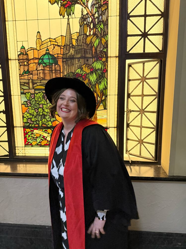 Woman smiling and wearing academic dress including an epic black Tudor bonnet with yellow tassel, black gown, and red stole. She is standing in front of a stained glass window of the Melbourne skyline.