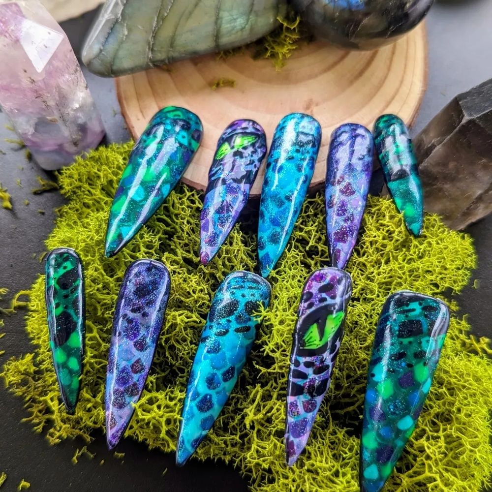 Long stiletto shaped nails with dragon scales in varying colors. Green, purple-blue, turquoise, and mixed green with blue. Each also has smaller black scales and two nails have one bright green dragon eye