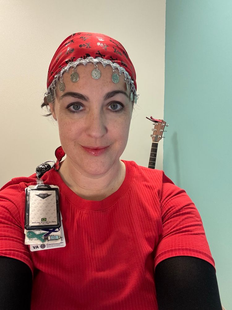 A Caucasian woman (me) with a red pirate bandana, red shirt, black sleeves and a guitar in the background in an office.