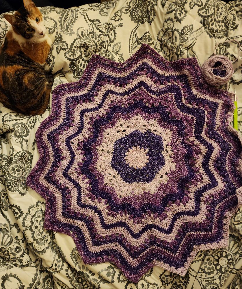 Another 14-point star blanket with three varying purple hues and a calico cat sitting nearby in the top left corner of the image. 