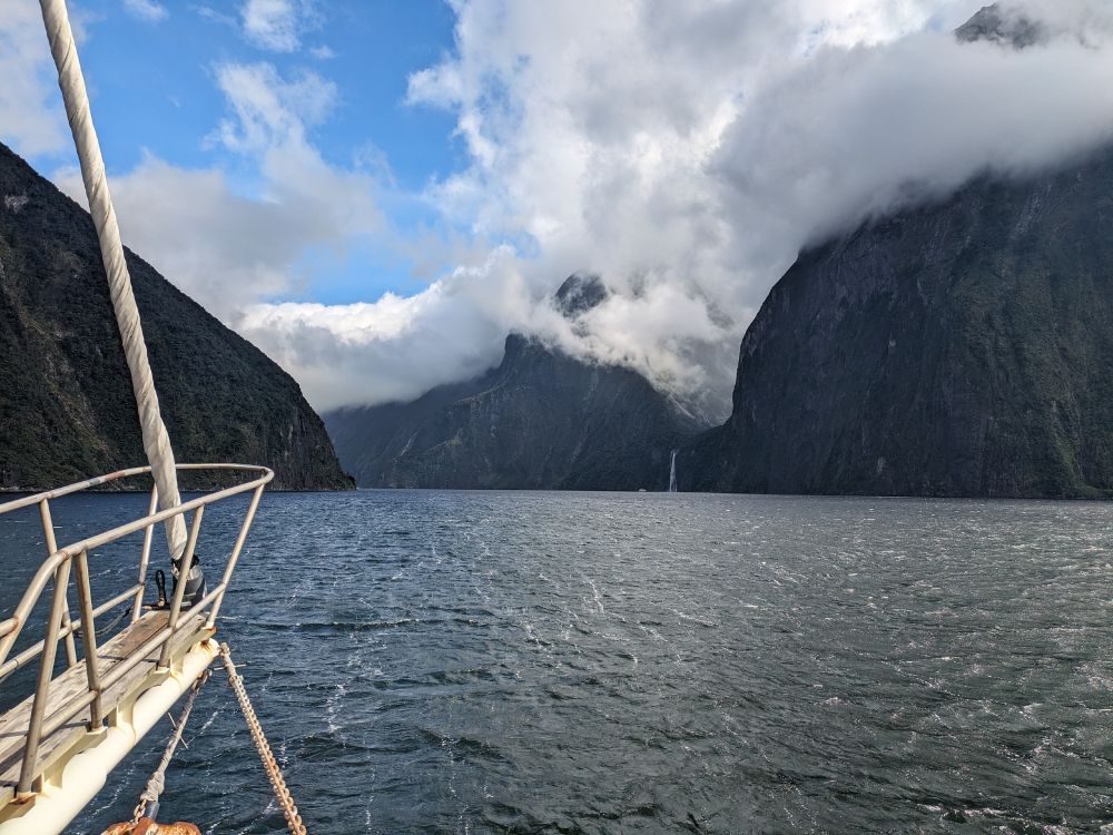 View from the bow of a boat travelling down a sound, with large cliffs on either side and clouds covering the cliffs on the right.