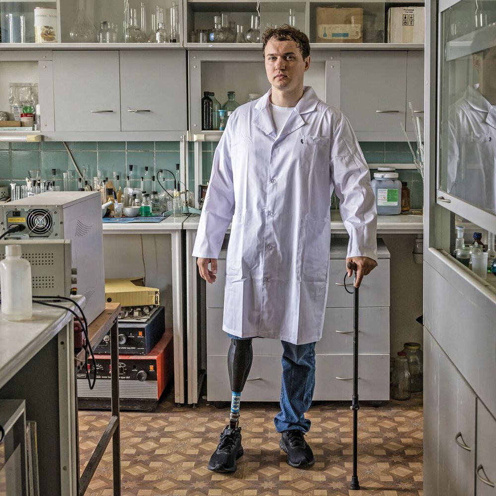 Artur Mylin wears a lab coat and stands for a portrait in an orderly lab space. He stands on one natural leg and one prosthetic leg and holds a cane. An array of flasks, some with stoppers and others without, are on the counter behind him and on the shelves above him. The brown, tiled floor alternates in a pattern of squares, diamonds, and eight-pointed stars.