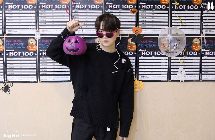 Yoongi is standing in front of a wall plastered with BB hot 100 charts topped by BTS and he’s wearing a casual black outfit and straight black hair reaching his eyelashes. He’s slid red sunglasses down his nose and he’s holding a purple Halloween pumpkin bucket next to his face.