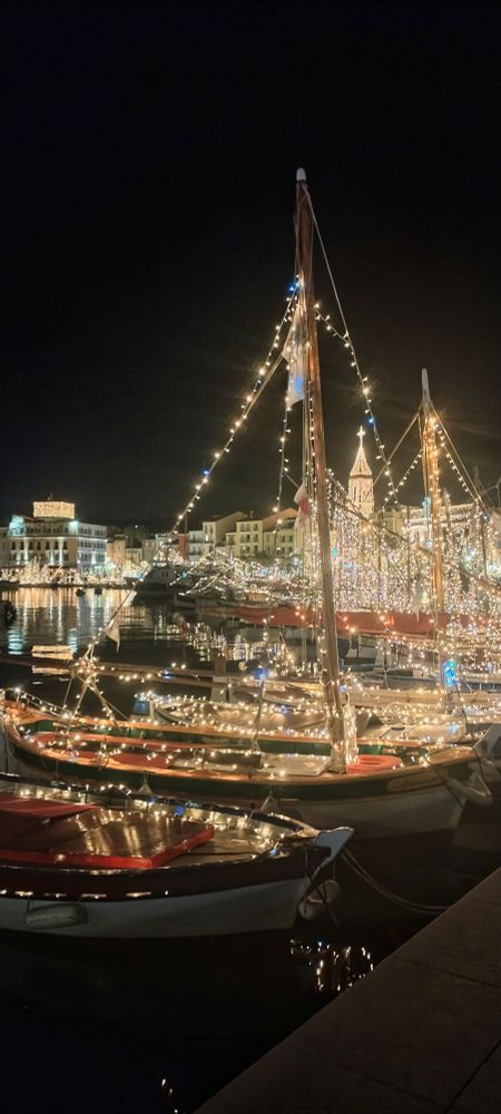 Sur le port envoie des pointus avec les mains illuminés avec des petites lampes et au fond on voit l'église et la tour de Sanary éclairés