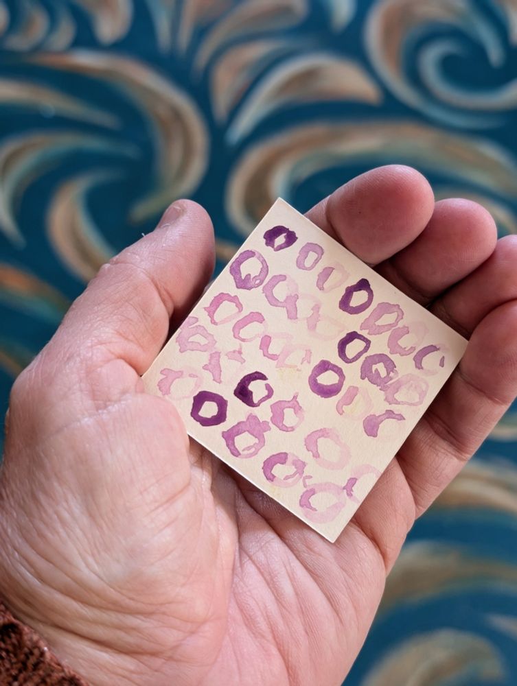 Held in the palm of the hand, a 2x2 inch paper  with  Varying shades of purple imperfect circles painted with water color 
