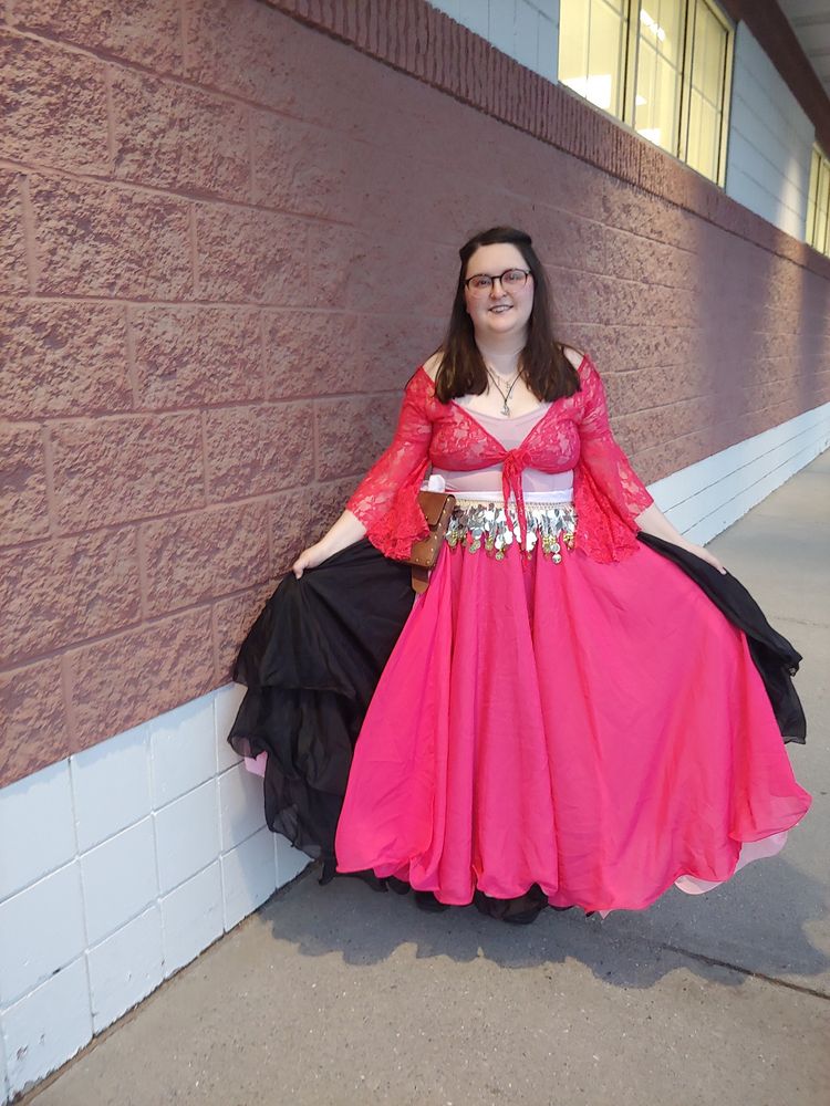 A woman in a pink and black dress with lacey sleeves. A white belt with silver coins sewn to it is around the waist.