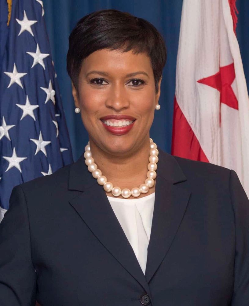Photo of Washington, DC Mayor Muriel Bowser. She is smiling, wearing a pearl necklace and earrings and dark suit in front of an American flag.