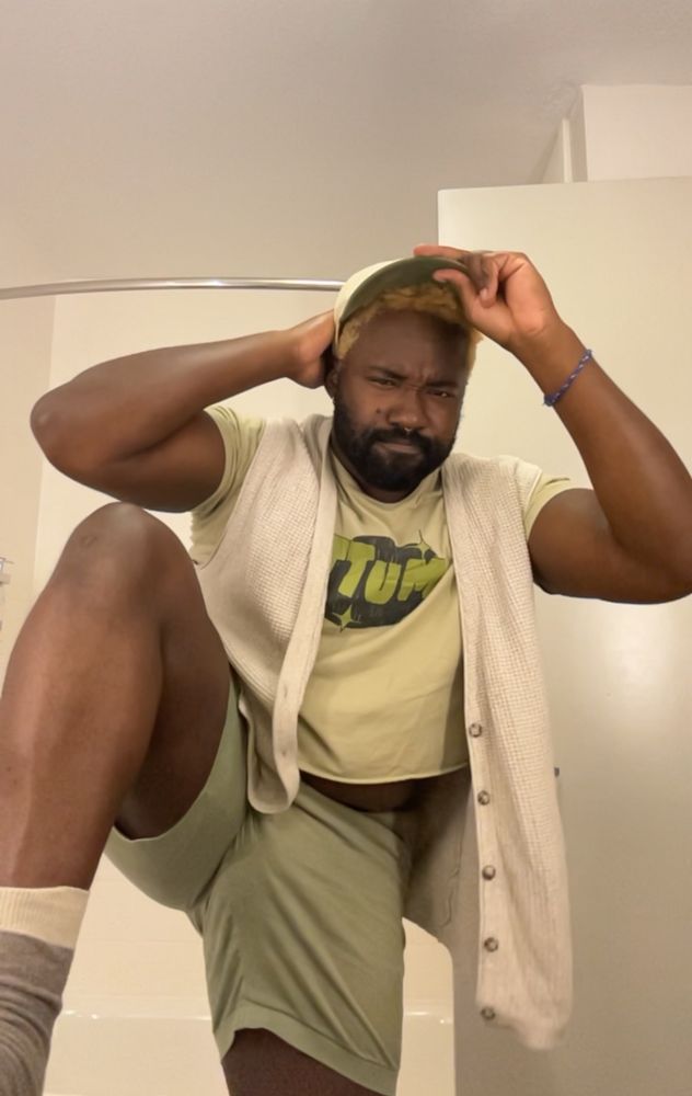 Thembospice selfie in a green outfit with a leg up. They’re wearing a crop top and shorts and a sleeveless sweater vest and a lil hat