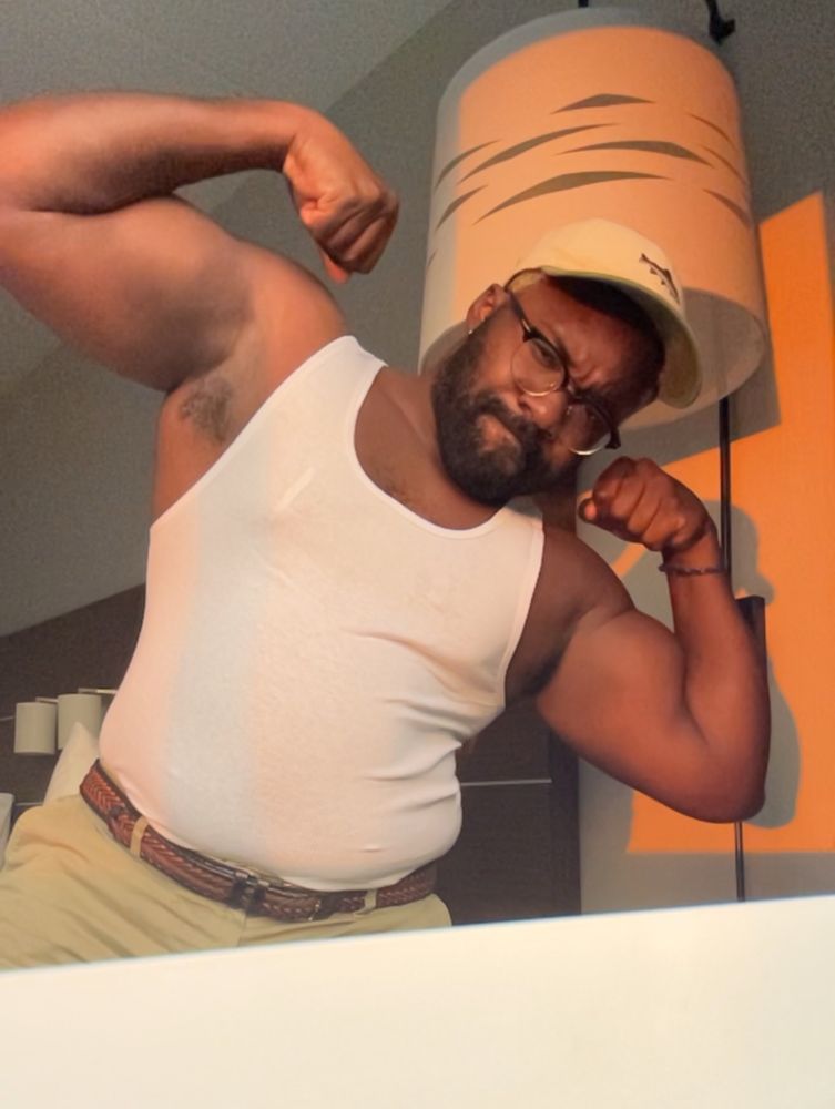 Thembospice double bicep flexing in a white tank, hat and green shorts