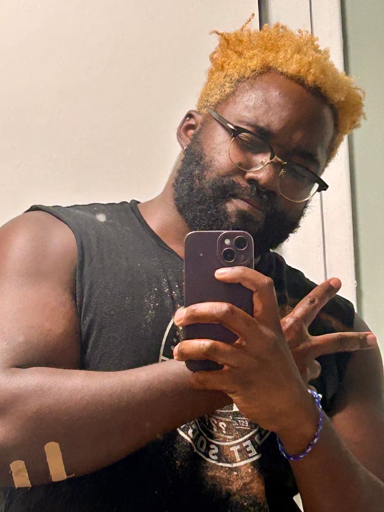 Thembo mirror selfie from the waist up with a black shirt, blonde hair and glasses throwing up a millennial ass peace sign 