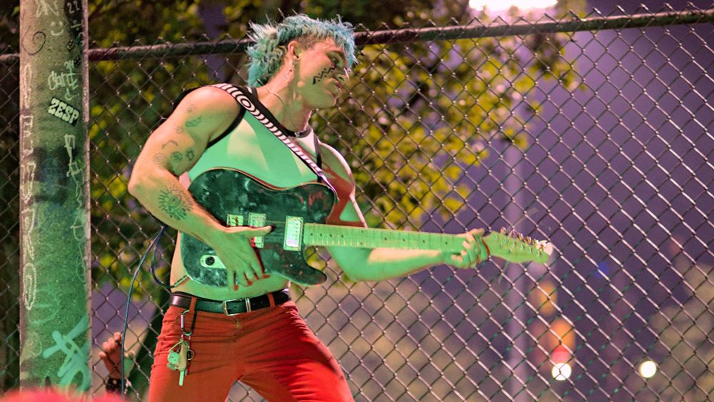 Photo of person playing guitar in front of skate park fence, face paint, blue hair