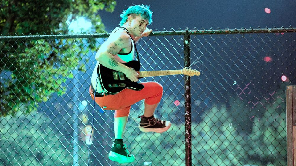 Photo of person climbing skate park fence while carrying guitar, face paint, blue hair