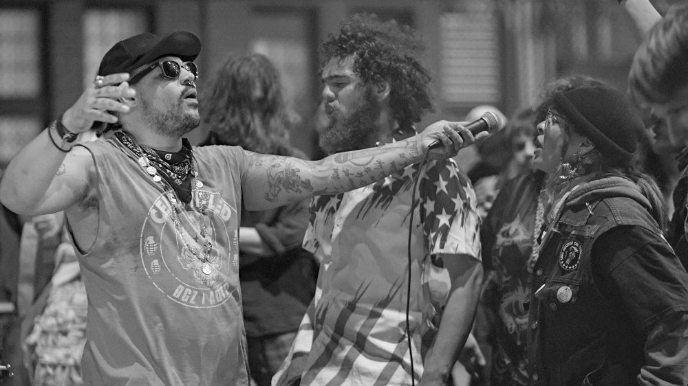 Monochrome photo of singer wearing sunglasses holding out microphone for person in crowd to sing into microphone, other concert attendees also seen