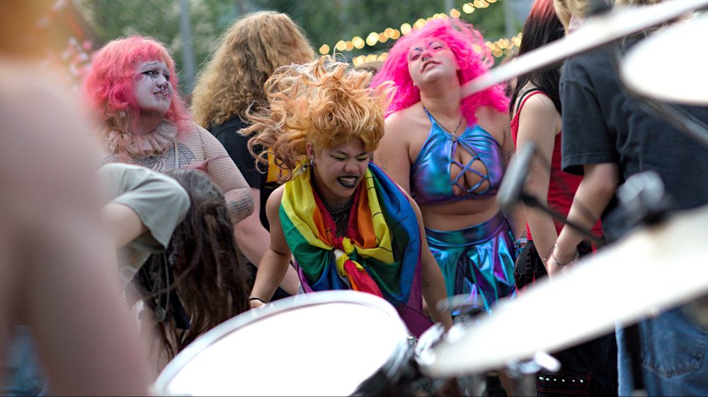 Photo of people dancing and gesturing to live music being played in front band playing, person in front wearing pride colored clothing, hair waving in the air, person behind wearing shiny blue clothing with pink hair, another person with red pink hair wearing white make up with face paintings
