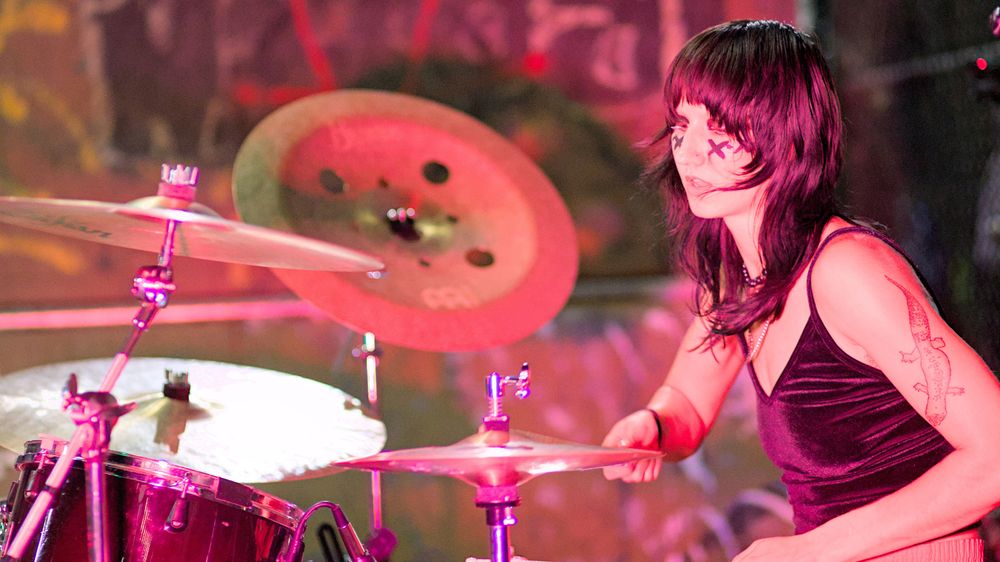 Photo of person with X's on their face playing drums for concert, drumming and percussion equipment seen
