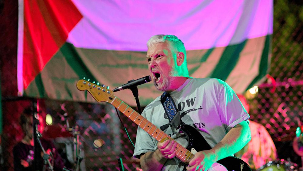 Photo of person singing into microphone, playing guitar and making facial expression, flag of Palestine and skate park fence in background