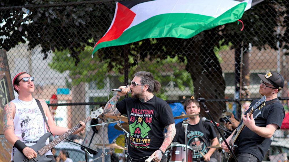 Photo of band playing, 4 people, left side person playing guitar, center person singing into microphone, background person playing drums, right side person playing guitar, flag of Palestine in background
