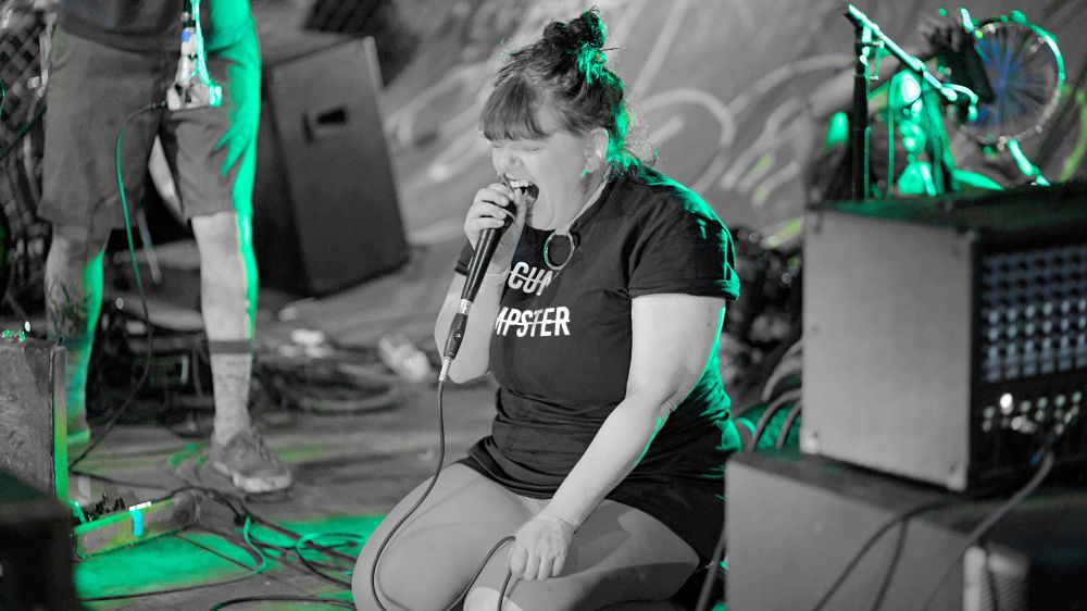 Mostly monochrome photo but with green highlights, person on knees singing into microphone, wearing a t-shirt that says see you emm dumpster, other concert equipment and skate park ramp seen in background.