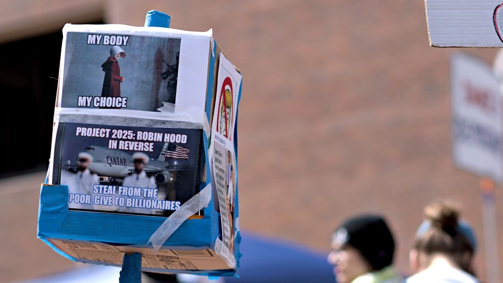 Protest sign, My body my choice with handmaid's tale picture, project 2025: robin hood in reverse, steal from the poor. give to the billionaires. Large Qatar jet picture with U.S. flag