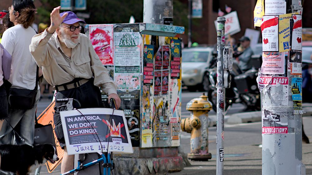 Person at the corner of Broadway and Pine waving with a protest sign, In america we don't do kings, they've defied our courts, deported Americans, disappeared people off the streets, attacked our civil rights, and slashed our services. The corruption has gone too far. No thrones. No crowns. No kings. No kings dot org.