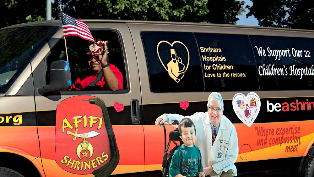 Photo of person driving black orange and yellow Shriners van, Shriners Hospitals for children love to the rescue. be a shriner, photo of young wheelchair user and doctor on side of van AFIFI Shriners