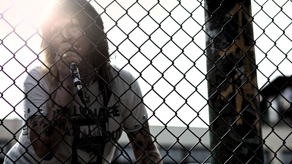 Photo of person with microphone singing behind skate park fence, sun shining from behind