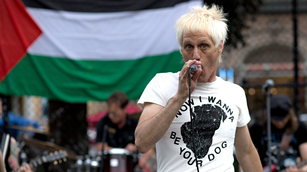 Photo of Steve E vocalist singer from the Taste Testors, band and large flag of Palestine in background. T-shirt Now i wanna be your dog, black dog pictured.