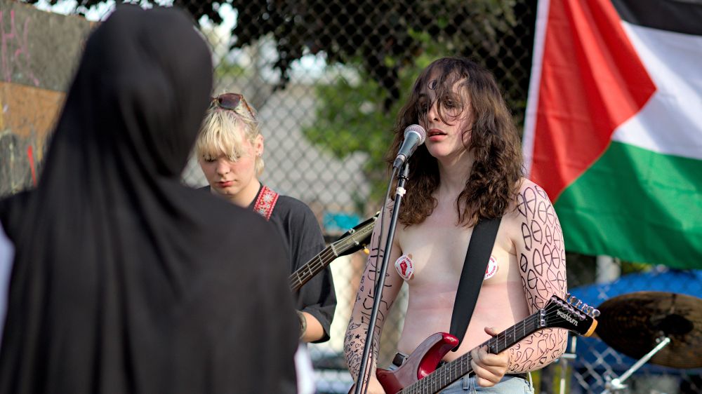 Photo of person playing guitar and singing, black pen drawings on both arms, no cooperation with u.s. fascism stickers on chest, some face paintings, another guitarist nearby, flag of Palestine in background