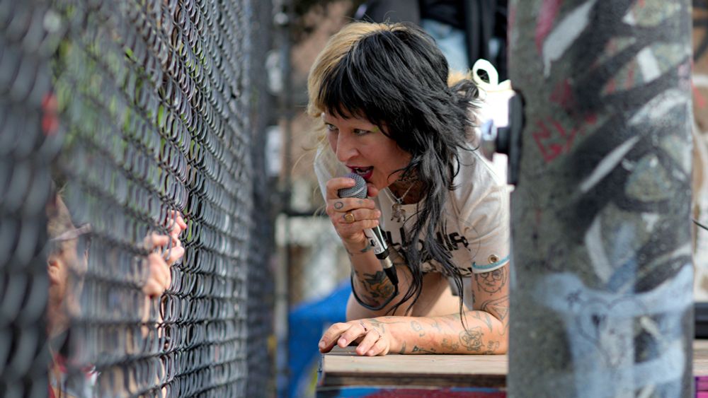 Photo of person with microphone kneeling down next to skate park fence, singing while looking through fence and people on other side of fence.