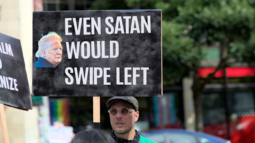 Person holding protest sign, even Satan would swipe left, photo of Donald Trump