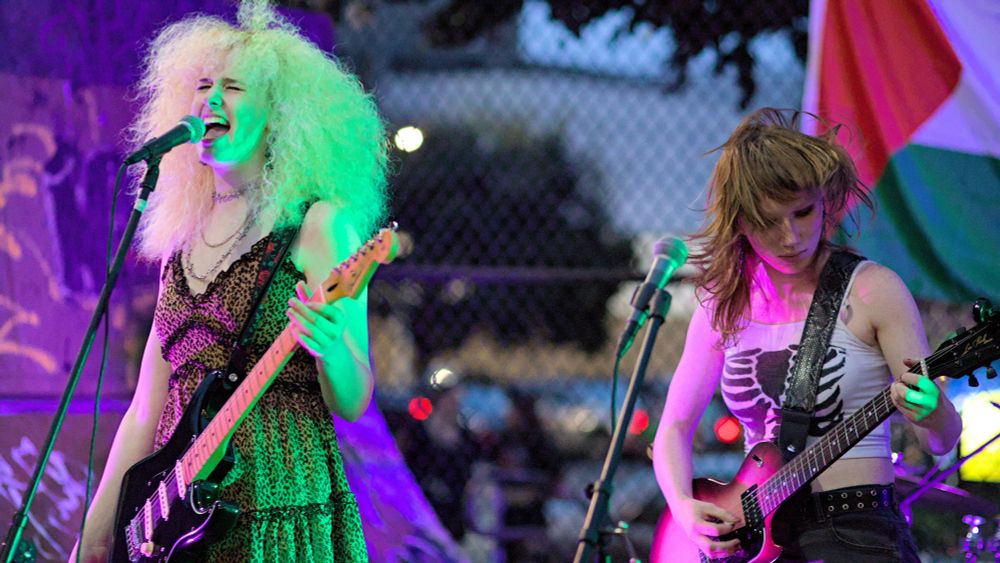 Two people on stage, one singing into microphone and playing guitar, another person also playing guitar