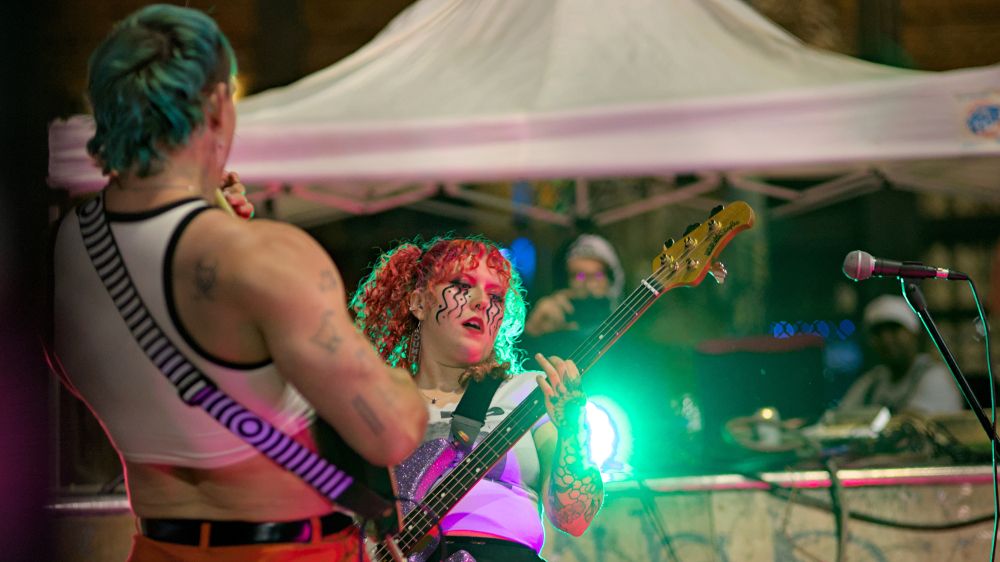 Photo of two people playing guitars in band, one person with multiple face paintings and red hair, green and red concert lights