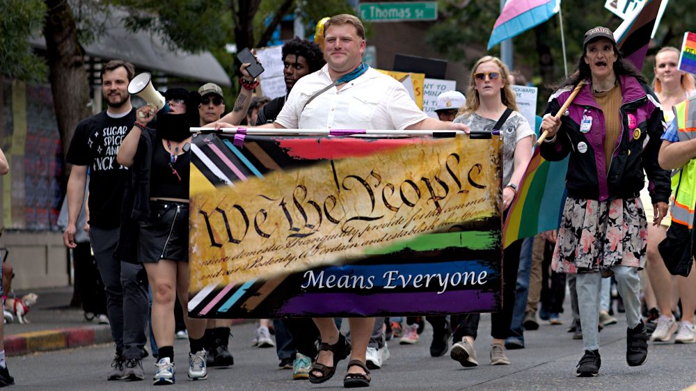 Photo of the front of the protest with a banner being held We the people means everyone, constitutional banner, multi inclusive colors on banner, protesters in background